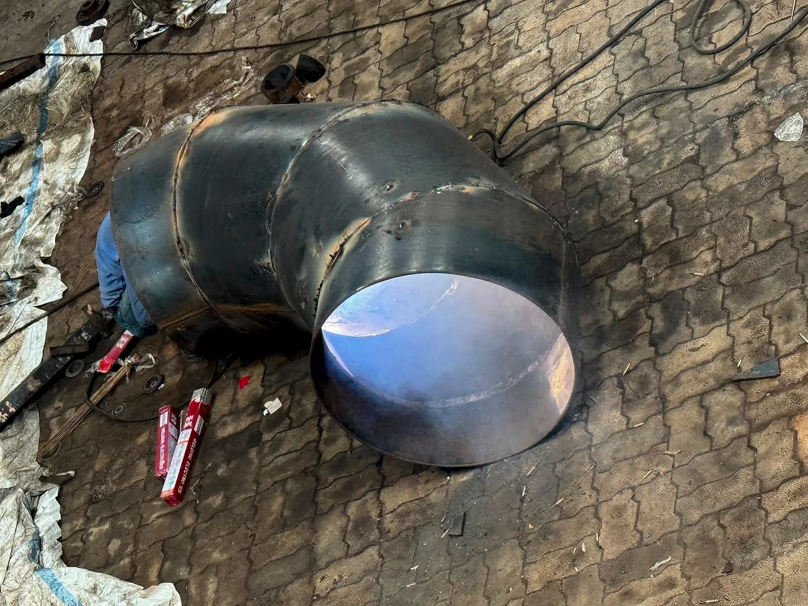 A large 90 degree mitre bend made of welded pipe segments resting on the floor